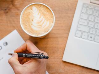 A hand holding a Ferguson Wellman branded pen next to a latte and laptop computer